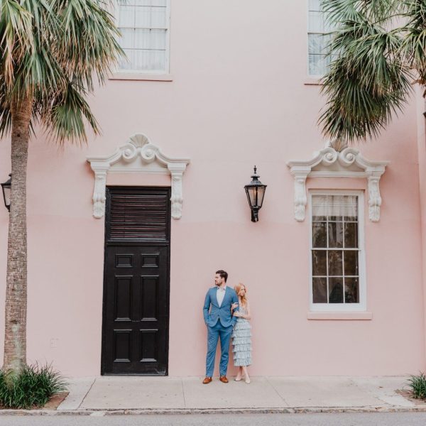 a man and woman standing on a sidewalk in front of a pink building in Downtown Charleston, SC. Photo By Bushel + Peck Photo